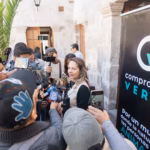 Mujer portavoz de Compromiso Verde sonríe ligeramente mientras habla con un grupo de reporteros que sostienen teléfonos inteligentes y micrófonos para grabar sus declaraciones.