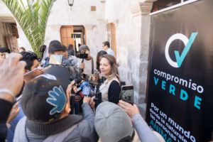 Mujer portavoz de Compromiso Verde sonríe ligeramente mientras habla con un grupo de reporteros que sostienen teléfonos inteligentes y micrófonos para grabar sus declaraciones.