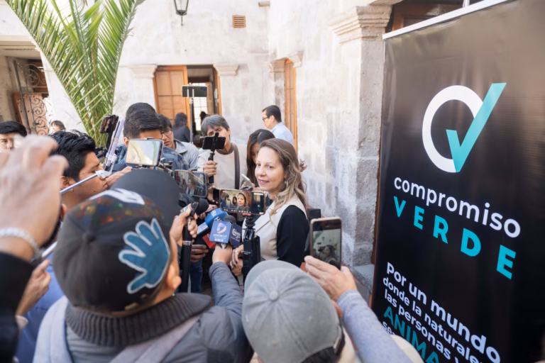 Mujer portavoz de Compromiso Verde sonríe ligeramente mientras habla con un grupo de reporteros que sostienen teléfonos inteligentes y micrófonos para grabar sus declaraciones.