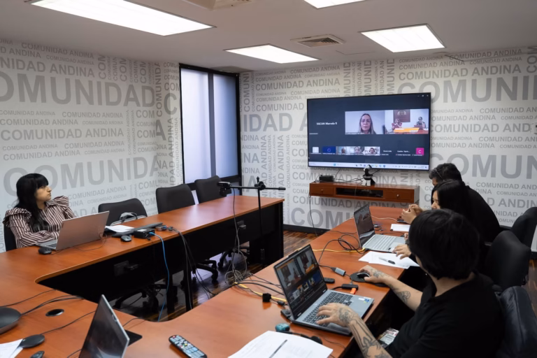 Funcionarios y equipo técnico reunidos alrededor de una mesa de conferencias con computadoras portátiles frente a una pantalla donde se transmite el webinar con el logo de la Comunidad Andina de fondo.